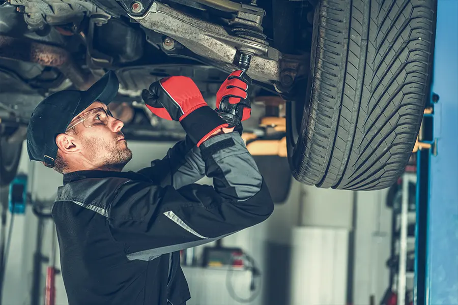 Mechanic in a workshop wearing a cap and red gloves, focused on tightening a bolt under a car's suspension. The setting conveys professionalism.