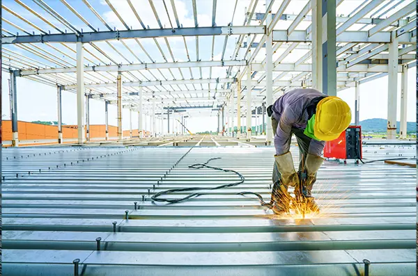 A construction worker in a yellow hard hat welds steel beams on a building site. Sparks fly as sunlight streams through the open framework, suggesting progress and energy.