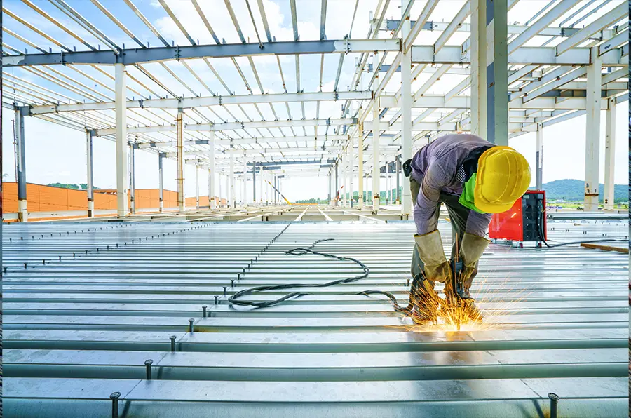 A construction worker in a yellow hard hat welds steel beams on a building site. Sparks fly as sunlight streams through the open framework, suggesting progress and energy.