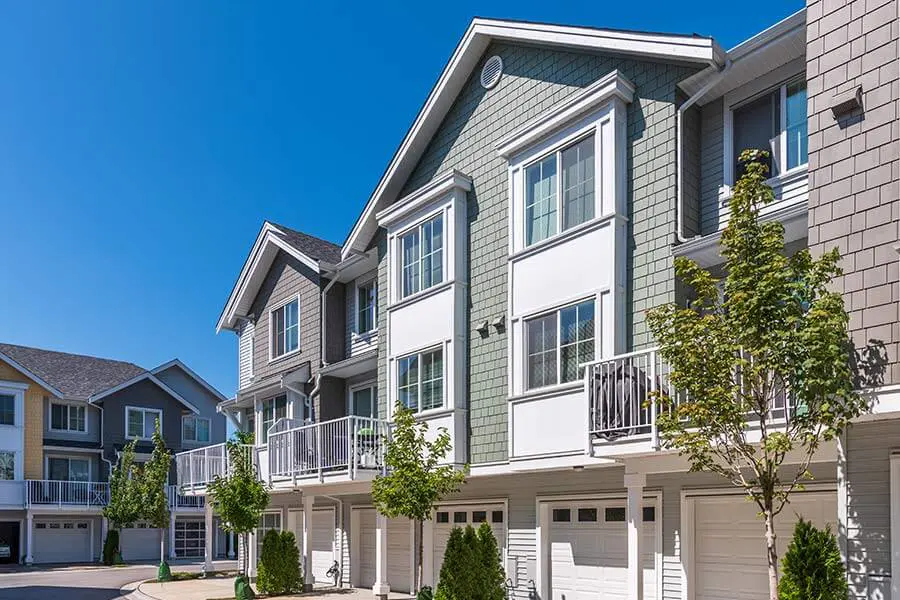 Modern townhouses with pitched roofs under a clear blue sky. The buildings feature large windows, balconies, and front garages, with small trees lining the street.