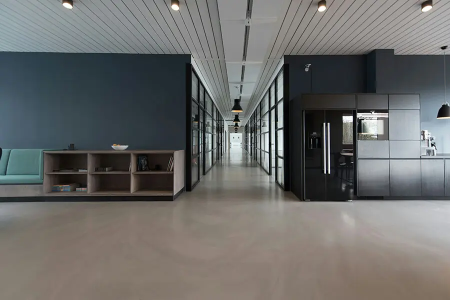 Modern office hallway with a sleek design, featuring glass-walled rooms, minimalist furniture, and overhead pendant lighting, creating a professional atmosphere.