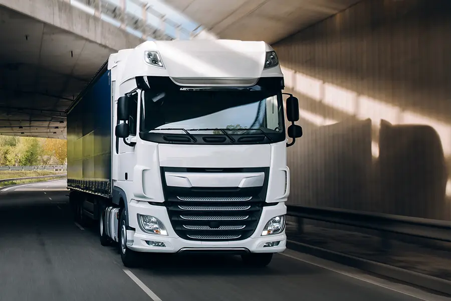 A large white semi-truck drives through a sunlit tunnel. The sleek vehicle conveys a sense of motion and reliability under clear skies.