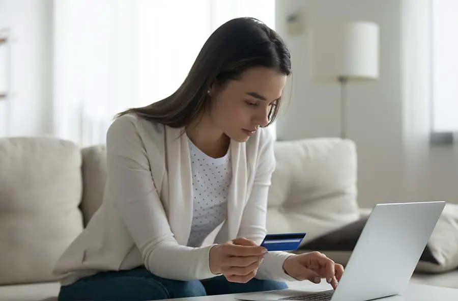 A woman sits on a couch, focused on her laptop while entering credit card details. The serene room reflects a neutral, soft tone.