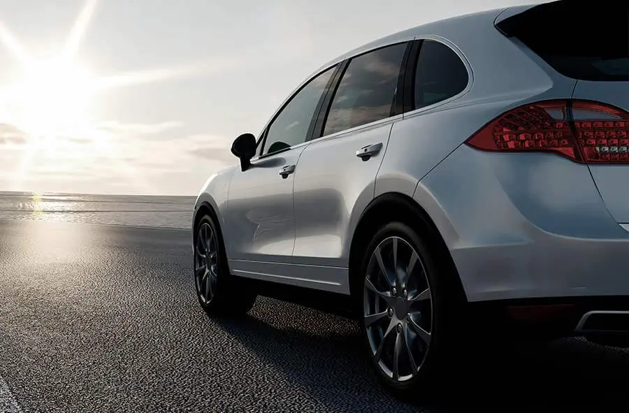 A sleek, silver SUV is parked on a coastal road at sunset. The sun casts a warm glow, creating a serene and peaceful atmosphere by the water.
