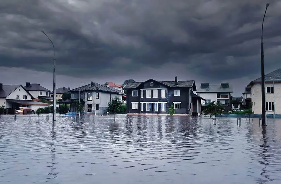 Flooded street with submerged road and houses under stormy skies. Dark clouds loom overhead, creating an ominous and tense atmosphere.