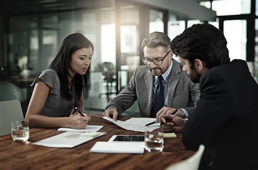 Three business professionals collaborate at a table with papers and a tablet. The setting is a modern office, conveying focus and teamwork.