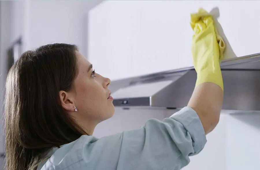 Woman in a light blue shirt and yellow rubber gloves wipes a kitchen exhaust hood with a cloth, focused expression. Clean, domestic setting.