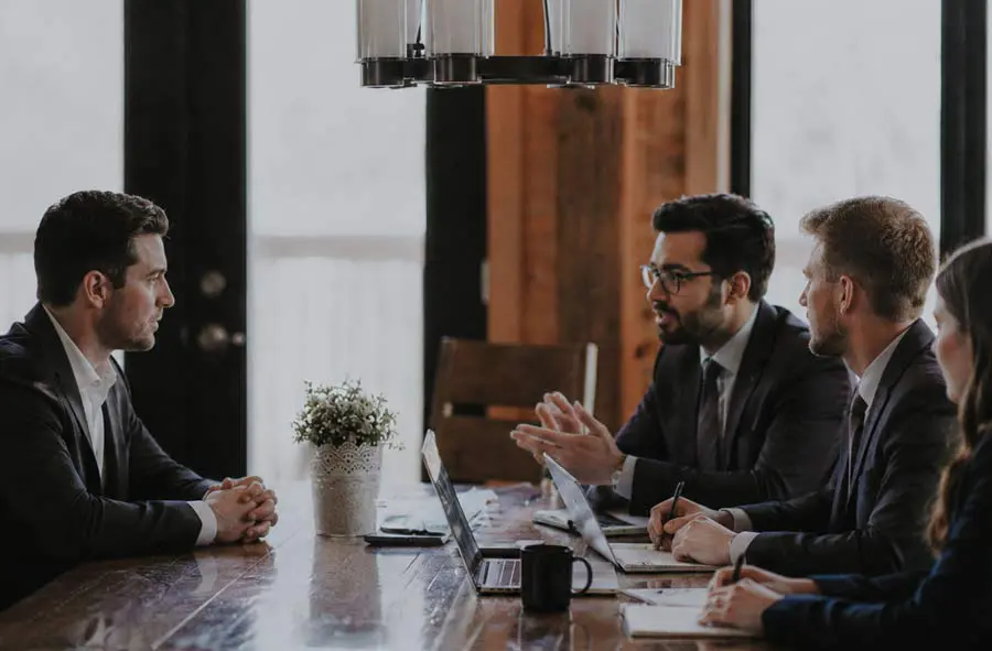 A group of four business professionals in a meeting around a wooden table. Two laptops are open, and one person is speaking, conveying focus.