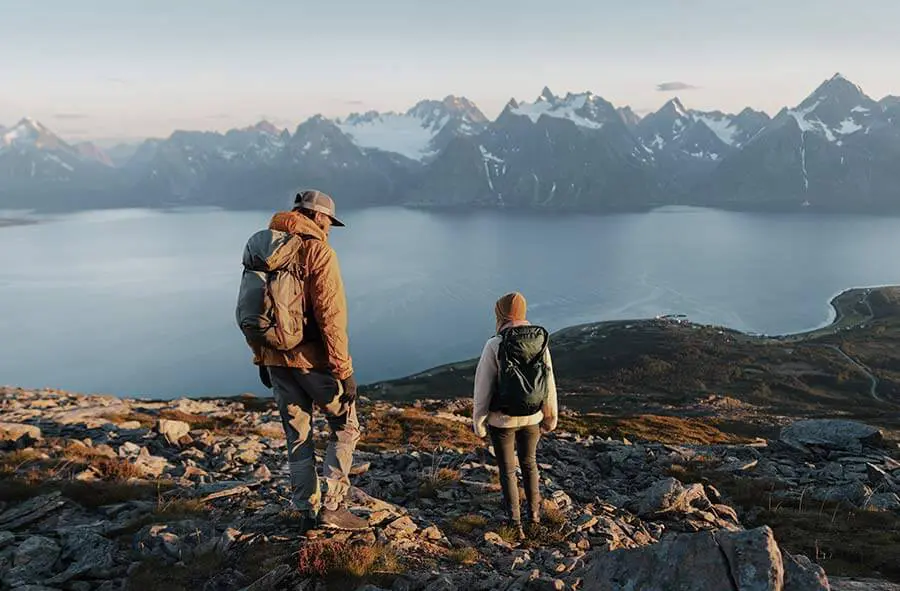 Two hikers with backpacks stand on a rocky hilltop overlooking a calm fjord and distant mountain range under a clear sky, suggesting tranquility and adventure.