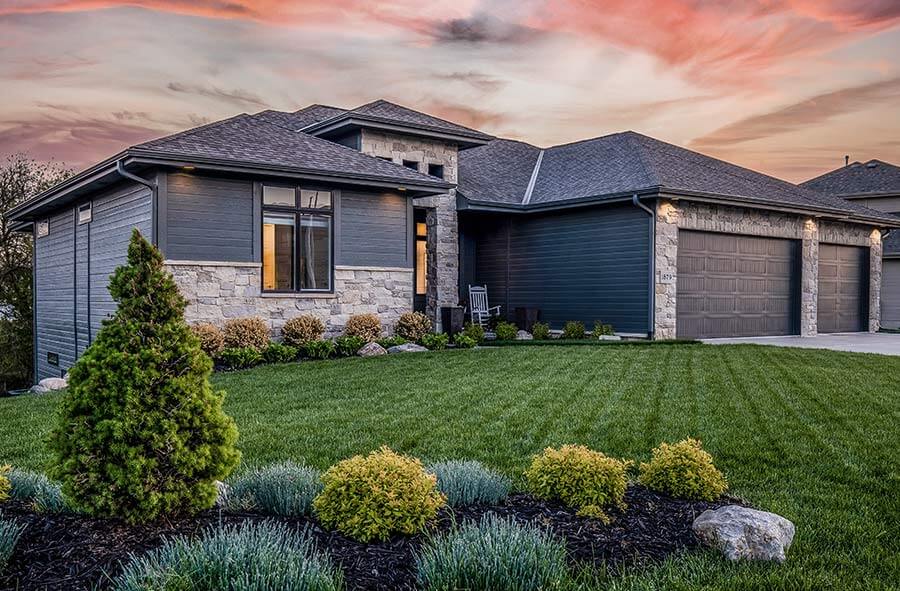 Modern house with gray siding and stone accents at sunset, surrounded by a manicured green lawn and shrubs. Warm light glows from the windows.